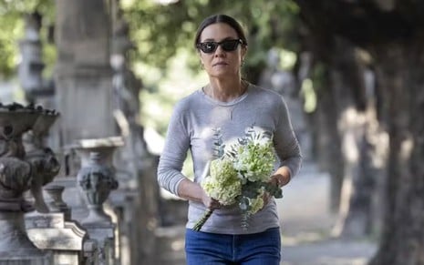 A mulher, de óculos escuros, veste camiseta cinza e calça jeans, caminhando em local arborizado, segurando um buquê de flores brancas e verdes.