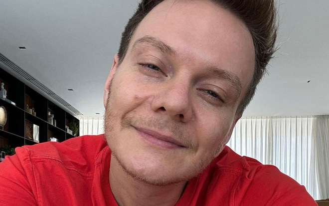 Michel Teló usa uma camiseta vermelha e dá um leve sorriso para a câmera