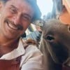 Um homem sorridente, usando camisa clara e avental, tira uma selfie ao lado de um burro. O fundo mostra paredes laranja e um pacote sobre uma superfície.