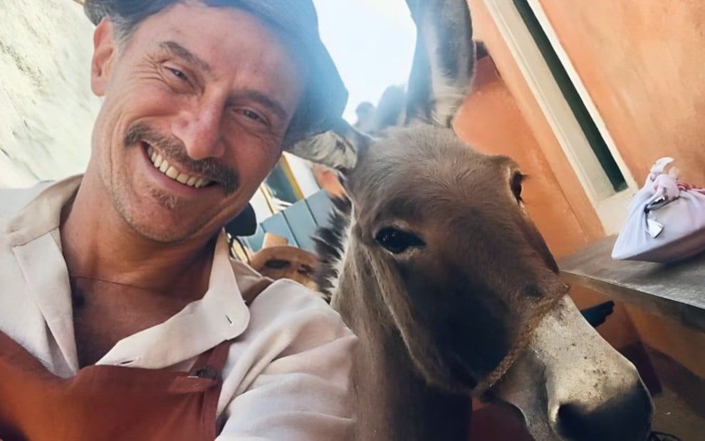 Um homem sorridente, usando camisa clara e avental, tira uma selfie ao lado de um burro. O fundo mostra paredes laranja e um pacote sobre uma superfície.