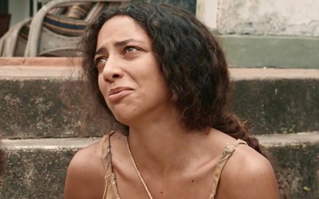 Alice Carvalho está sentada e faz uma expressão de choro em cena da novela Renascer como a personagem Joana