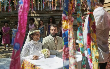 Duda Santos e Humberto Carrão estão ajoelhados na frente do altar; ela usa um vestido de noiva com véu preso na cabeça, e ele, um terno cinza