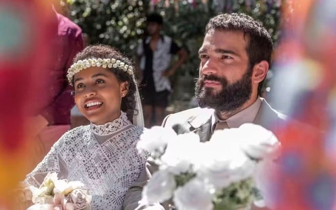 Humberto Carrão e Duda Santos estão parados no altar e sorriem para Chico Diaz, intérprete do padre Santo --fora do quadro-- em cena de Renascer