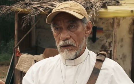 Chico Diaz caracterizado como padre Santo; ele usa batina branca e boné e está tenso em cena de Renascer