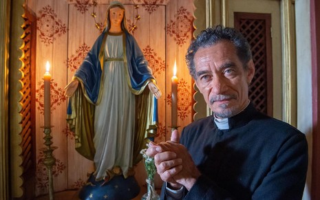 Chico Diaz está caracterizado como seu novo personagem, padre Santo do remake de Renascer, e posa perto de uma imagem de Nossa Senhora