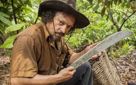 O ator Fabio Lago usa chapéu e camisa surrada e está com um facão nas mãos como o personagem Venâncio de Renascer