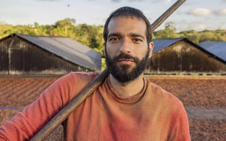 O ator Humberto Carrão segura uma enxada e está em ambiente de fazenda no remake de Renascer, da Globo