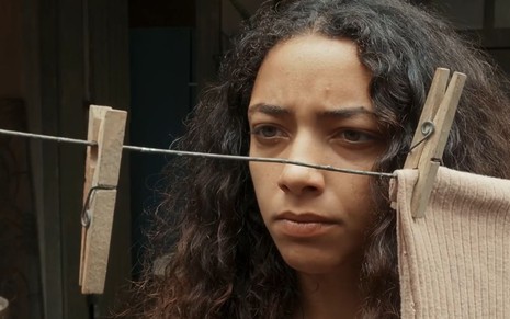 Alice Carvalho caracterizada como Joana; ela está séria, por trás do varal vazio, em cena de Renascer