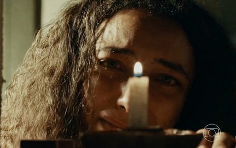Alice Carvalho está em close, com semblante entristecido e diante de uma vela, em cena da novela Renascer como Joana