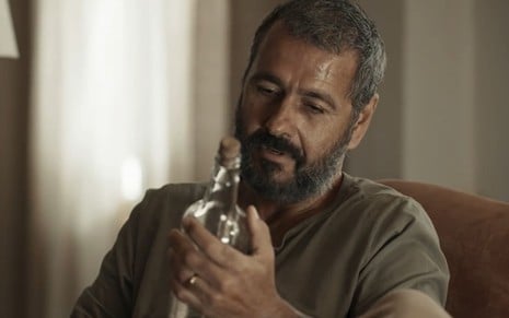 Marcos Palmeira caracterizado como José Inocêncio; ele usa uma camiseta cinza e está de perfil para a câmera, enquanto segura a garrafa com o diabinho