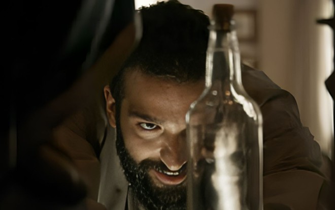 Humberto Carrão como José Inocêncio aproxima o rosto de uma garrafa, em que se vê um diabo de costas, em cena de Renascer