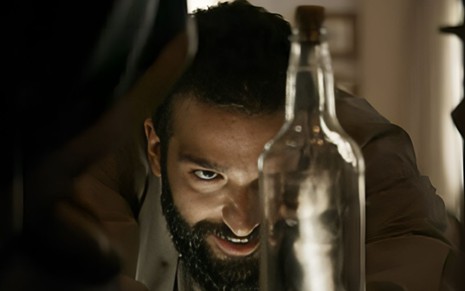 Humberto Carrão como José Inocêncio aproxima o rosto de uma garrafa, em que se vê um diabo de costas, em cena de Renascer