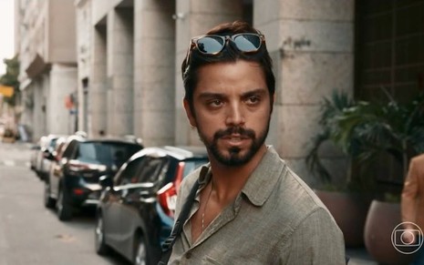 Rodrigo Simas em cena de Renascer caracterizado como José Venâncio; ator está em uma rua com óculos escuros na cabeça