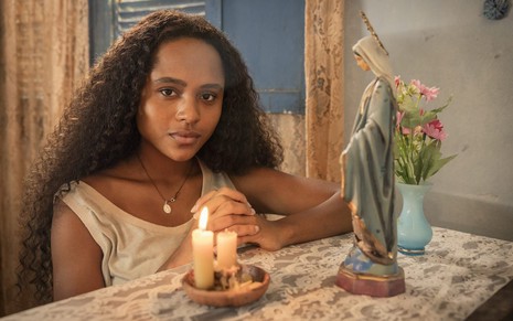 A atriz Duda Santos está diante de uma vela acesa e de uma imagem de santa como a personagem Maria Santa de Renascer