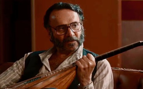 O ator Almir Sater segura instrumento musical e olha para o lado como Rachid na novela Renascer, da Globo