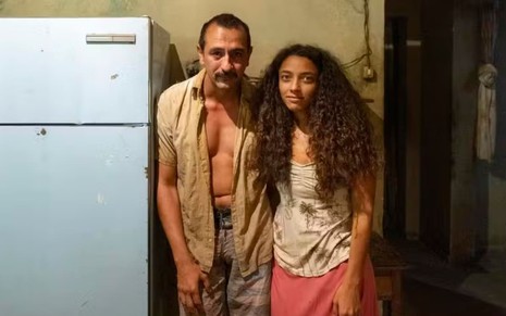 Irandhir Santos, o Tião Galinha, usa uma camisa de botão surrada e uma bermuda; Alice Carvalho usa blusa e saia surradas em cena de Renascer