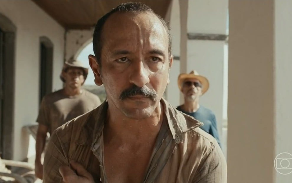 O ator Irandhir Santos está em destaque em cena de Renascer como Tião Galinha, outros dois atores aparecem ao fundo desfocados
