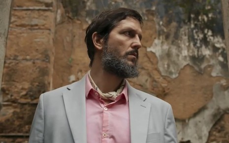 Vladimir Brichta caracterizado como Egídio; ator usa um terno cinza e camisa rosa e está de perfil; ele exprime tensão enquanto olha para o lado esquerdo em cena da novela Renascer