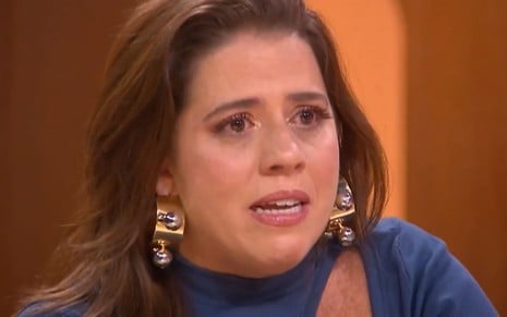 Tati Machado está com os olhos marejados; ela usa uma blusa azul e brincos de argola dourados. O semblante está deformado pelo esforço das lágrimas no Saia Justa