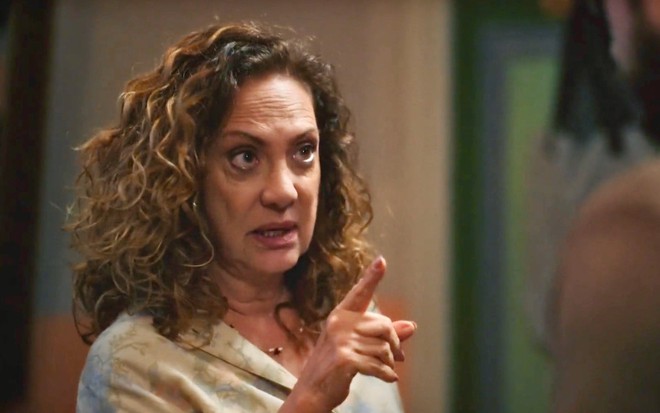 Em cena de Terra e Paixão, Eliane Giardini está conversando com alguém; ela usa blusa caramelo