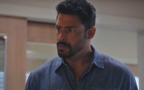 Cauã Reymond caracterizado como Caio; ele usa uma camisa azul e encara Tony Ramos, fora do quadro, com o semblante exalando fúria em cena de Terra e Paixão