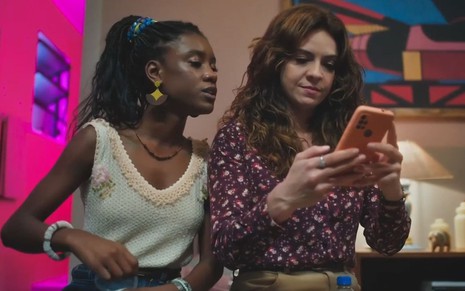 Em cena de Terra e Paixão, Camilla Damião está olhando o celular de Renata Gaspar por cima do ombro da atriz