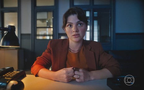 Petra (Debora Ozório) sentada diante de uma mesa com roupa vermelha em cena da novela Terra e Paixão