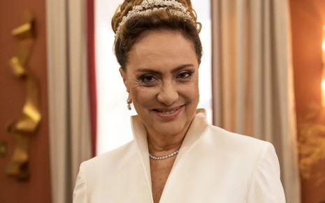 Eliane Giardini caracterizada como Agatha; atriz está com os cabelos presos num coque alto e usa um vestido de noiva creme, liso e com gola alta
