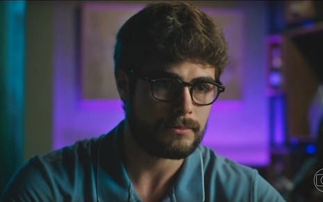 Rafael Vitti usa óculos e camisa azul e tem olhar triste em cena da novela Terra e Paixão como Hélio