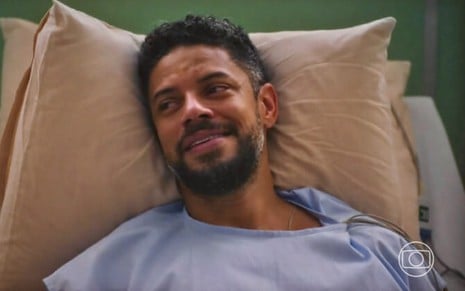 Paulo Lessa usa avental de paciente e está deitado em um leito em cena da novela Terra e Paixão, da Globo