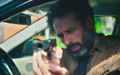 O ator César Ferrario está apontando uma arma de dentro de um carro em cena da novela Terra e Paixão na qual faz um matador