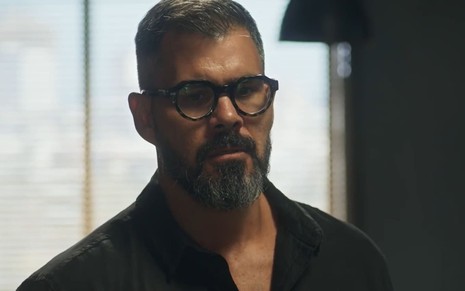 Juliano Cazarré caracterizado como Pascoal; ele usa uma camisa preta e óculos de aro preto em cena de Fuzuê. O semblante exprime confusão.
