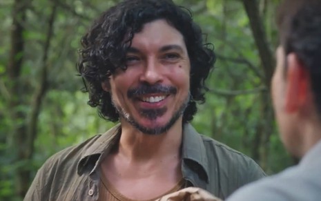 Paulo Roque caracterizado como Sidney; ele usa uma camisa verde parcialmente aberta e dá um sorriso perturbador em cena de Terra e Paixão