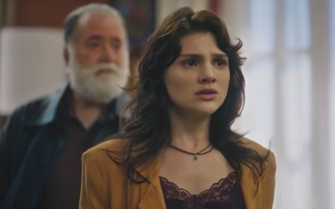 Debora Ozório caracterizada como Petra; ela encara um ponto fixo com o semblante chocado em cena de Terra e Paixão. Ao fundo, desfocado, está Tony Ramos, o Antônio da novela