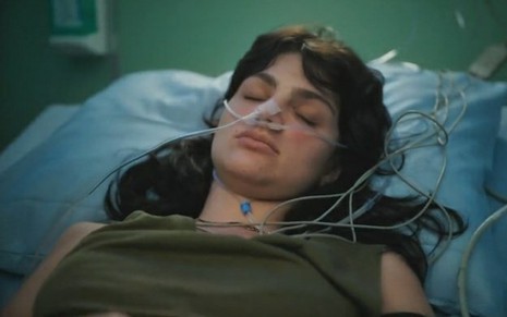 A atriz Debora Ozório está em um leito com acesso no nariz e com os olhos fechados em cena da novela Terra e Paixão