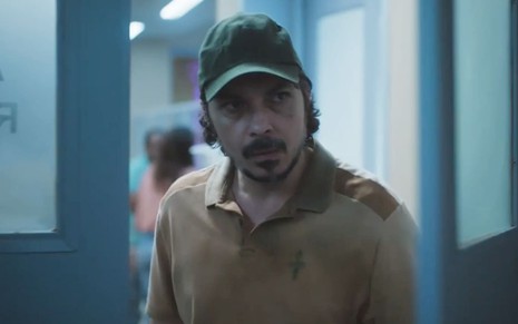 Paulo Roque caracterizado como Sidney em cena de Terra e Paixão; ele usa uma camiseta bege surrada e boné preto. O semblante exprime preocupação.