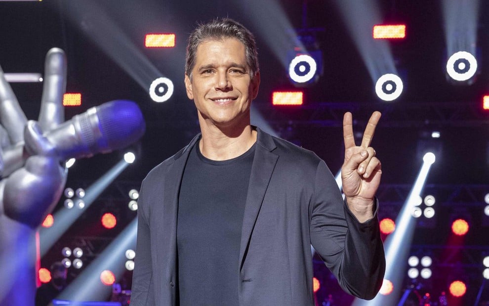 Marcio Garcia faz um V com os dedos, no cenário do The Voice Kids
