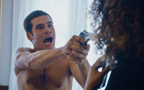 Nicolas Prattes está sem camisa e aponta uma arma para Barbara Reis em cena da novela Todas as Flores, da Globo