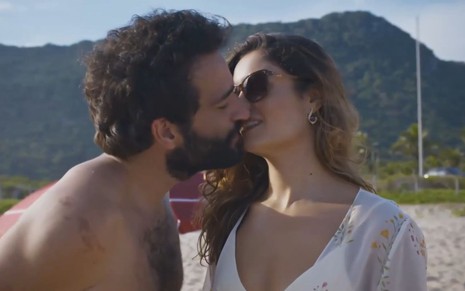 Na praia, Humberto Carrão e Sophie Charlotte se beijam; ele está sem camisa, ela usa óculos escuros