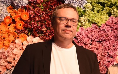 De blaxer preto e blusa branca, João Emanuel Carneiro posa em fundo cheio de flores coloridas