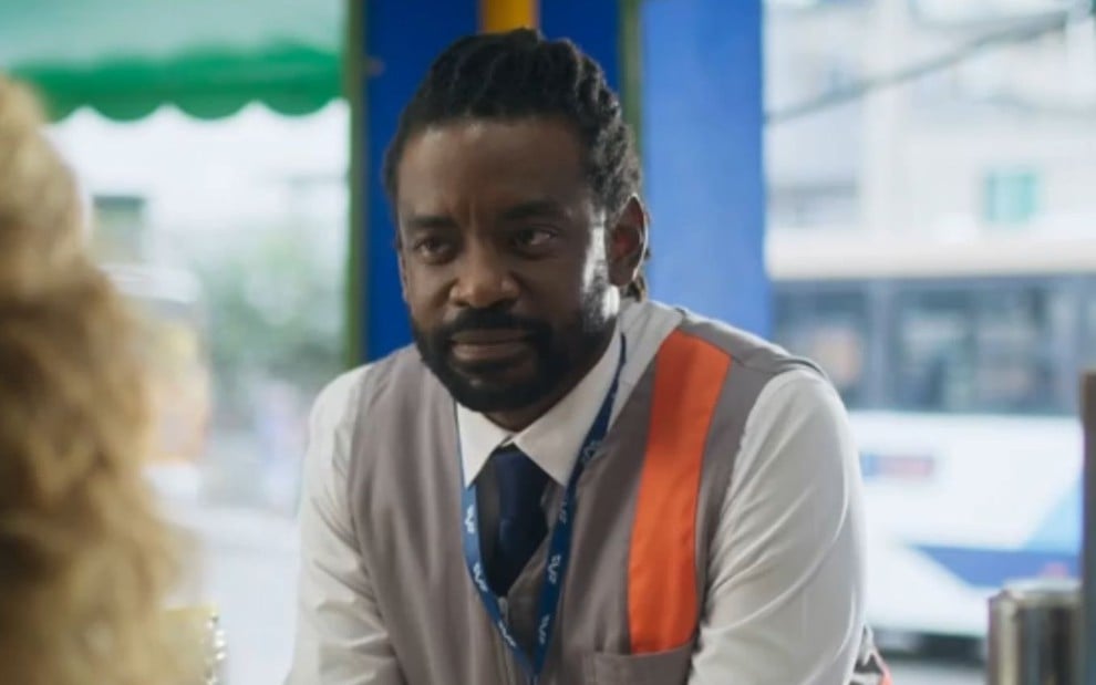 O ator Fabrício Boliveira caracterizado como Jão com o uniforme de fiscal de ônibus em um terminal em cena de Volta por Cima