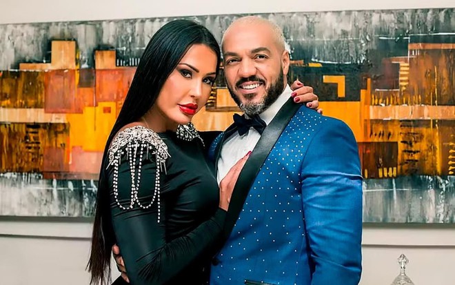 Gracyanne Barbosa usa um vestido preto e abraça Belo, que está com um terno azul; os dois encaram a câmera