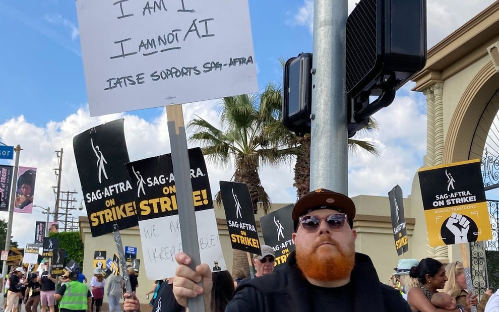 Um homem ruivo, com roupas pretas, segura uma placa com os dizeres "eu sou eu, eu não sou inteligência artificial" em piquete da greve dos atores