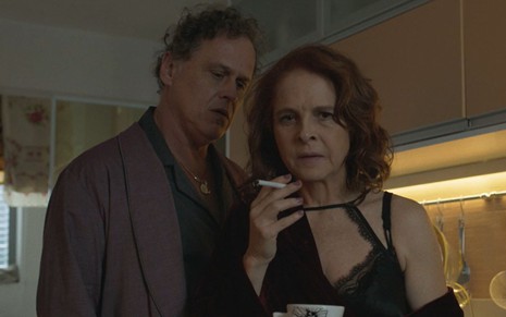 Guilherme Fontes está em pé ao lado de Drica Moraes, que segura um cigarro e uma xícara, em cena da série Os Outros
