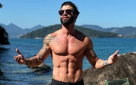 Gusttavo Lima está sem camisa, de braços abertos e usa óculos de sol; ao fundo é possível ver o mar e uma ilha