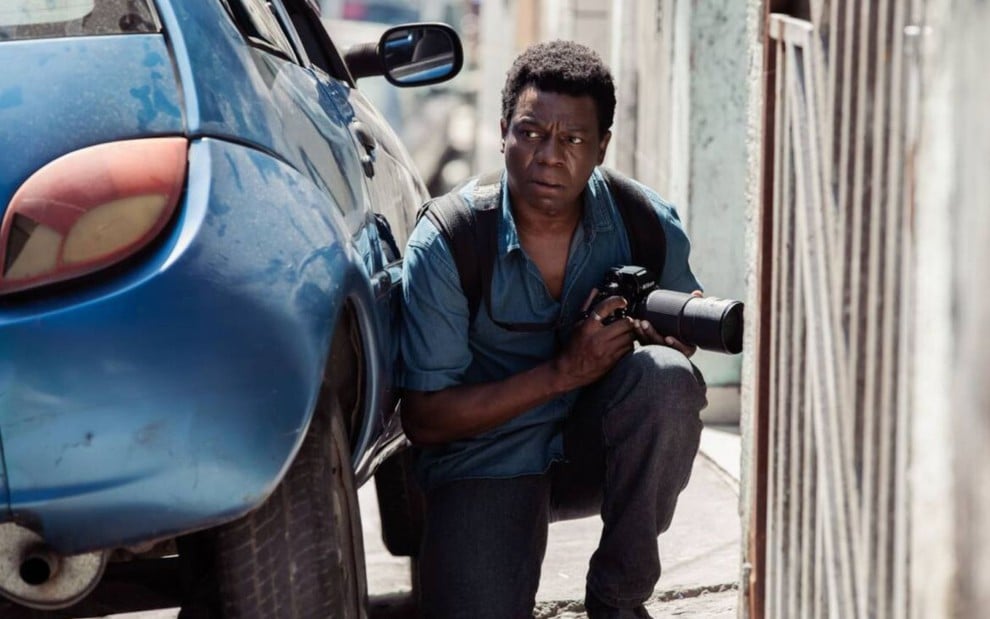 Alexandre Rodrigues segura uma câmera fotográfica e se esconde atrás de um carro em cena de Cidade de Deus: A Luta Não Para