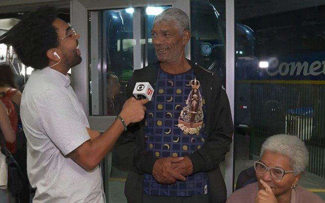 Repórter Henrique Silva dá risada e segura microfone enquanto fala com Carlos Alberto; Júlia está sentada no canto direito da imagem