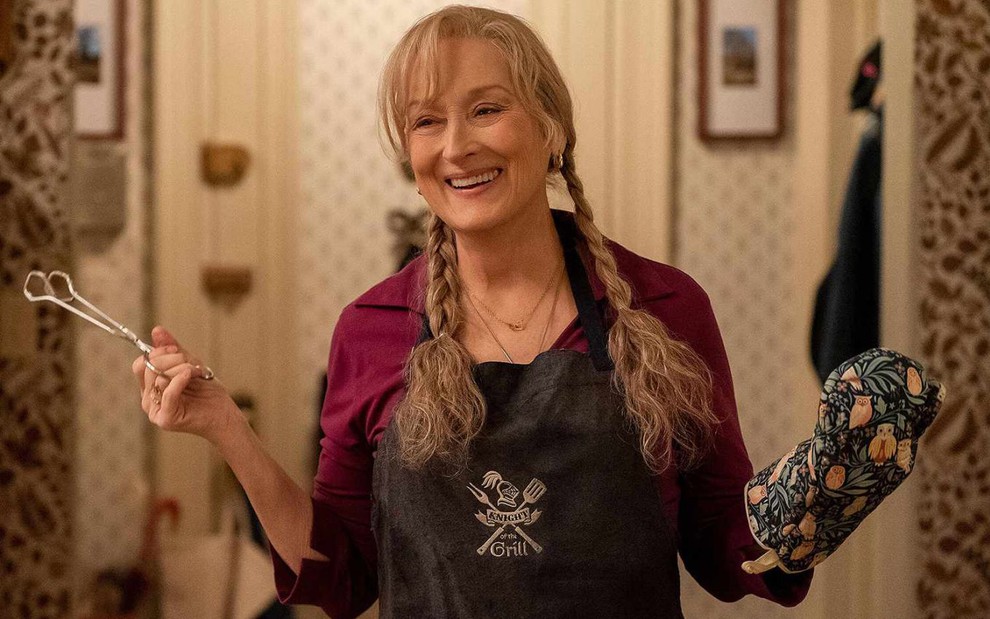 Meryl Streep sorri na cozinha enquanto segura uma pinça e usa uma luva de forno e um avental