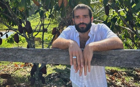 Humberto Carrão posa com uma blusa branca; ele está com os braços apoiados em uma cerca