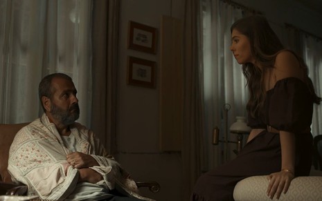 Em cena de Renascer, Marcos Palmeira está sentado em sua cadeira enrolado em uma manta e Gabriela Medeiros está em frente a ele